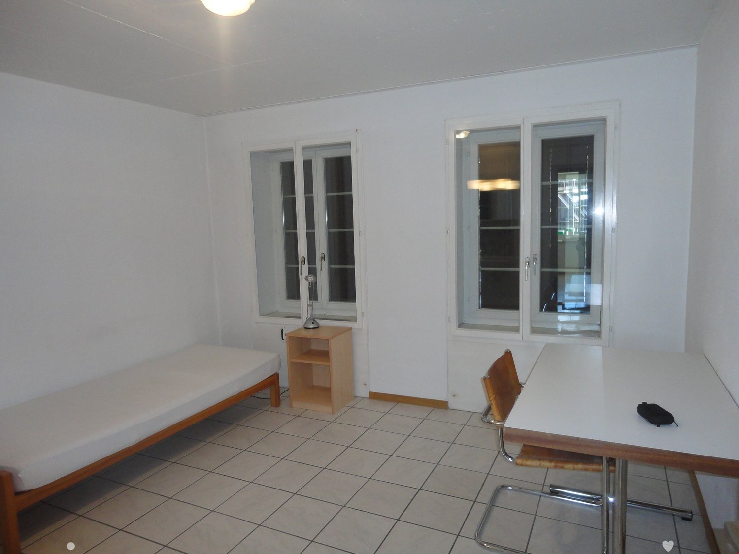 small room with single bed, wooden table, desk, wooden chair, lamp on table, white walls, windows, white tile floor