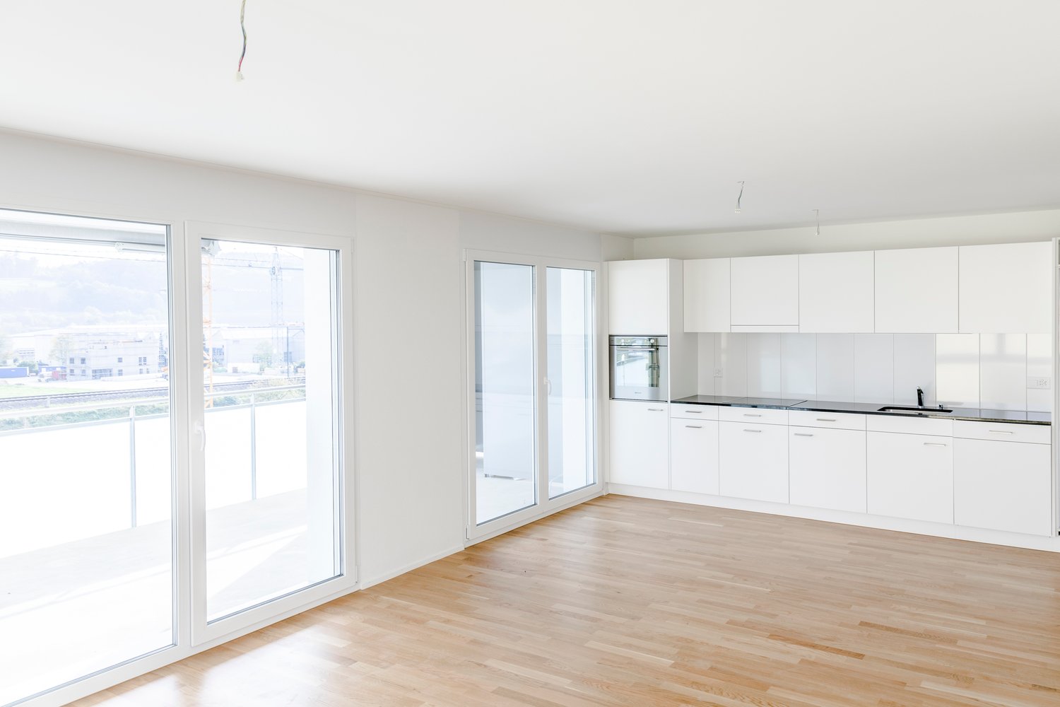 spacious room, white walls, white kitchen cabinets, wooden floor, double sliding doors leading to a balcony