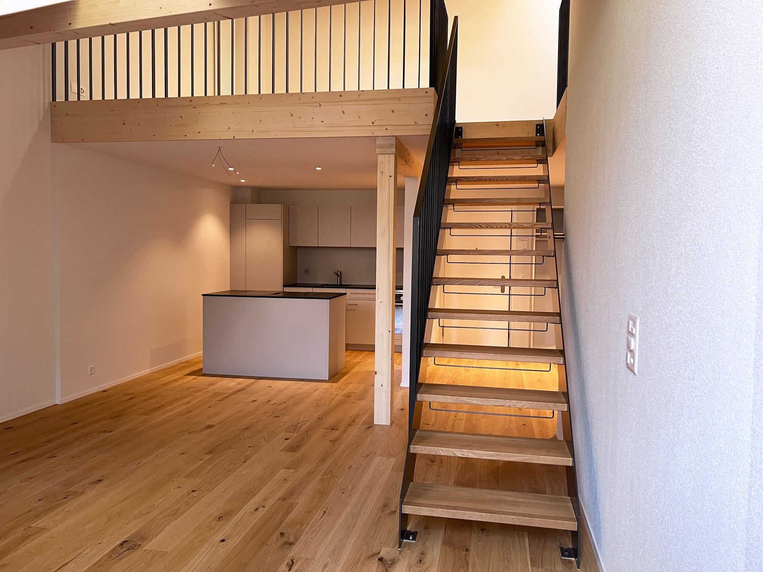 Open-concept living space with hardwood floors, a kitchen island, and a wooden staircase leading to a loft or upper level.