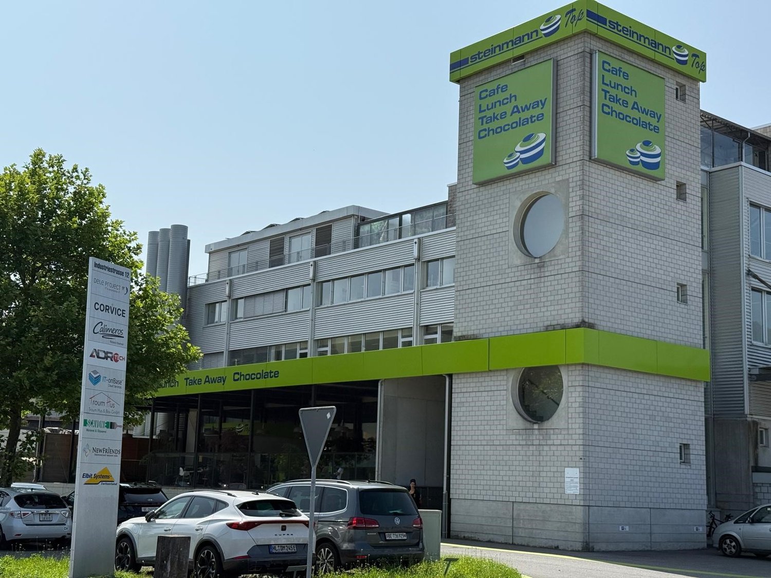 A multi-story commercial building with a Steinmann cafe and restaurant on the ground floor. The building has a large green and white sign advertising their cafe, lunch, take away, and chocolate offerings. There are several cars parked in front of the buil