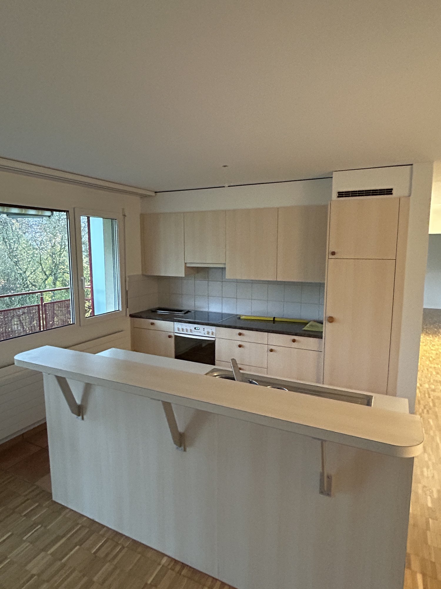 Contemporary open kitchen with wooden floors, large island counter, modern cabinets, built-in oven, gas stove, central sink, and windows with a view.