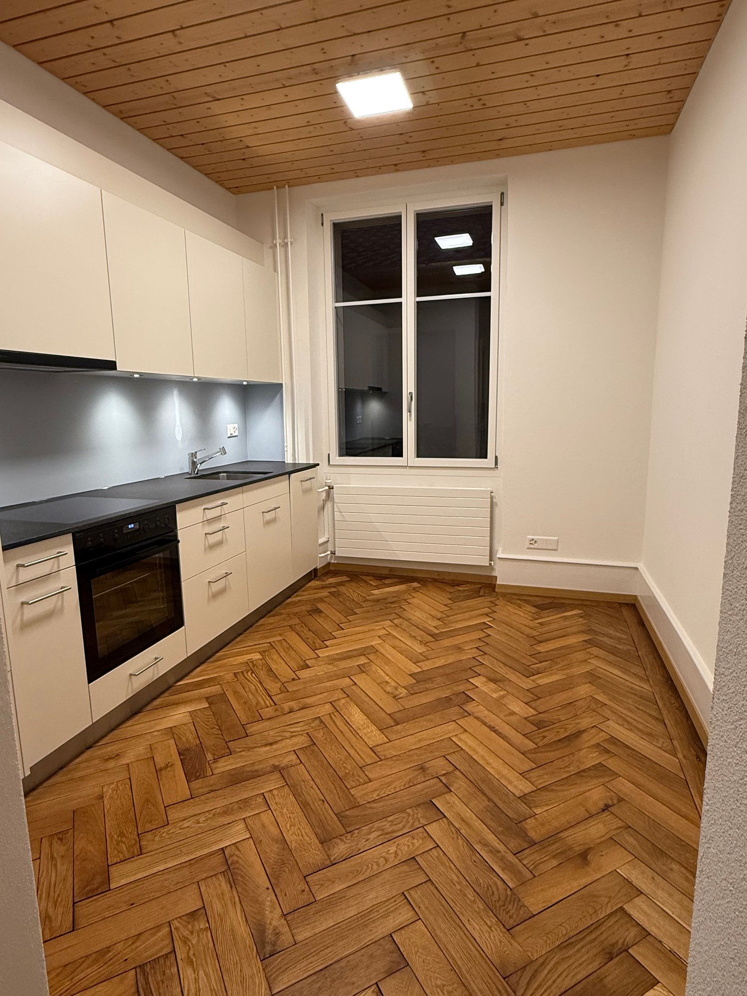 modern kitchen, wooden herringbone flooring, white cabinets, black countertop, integrated appliances, large window, ceiling light