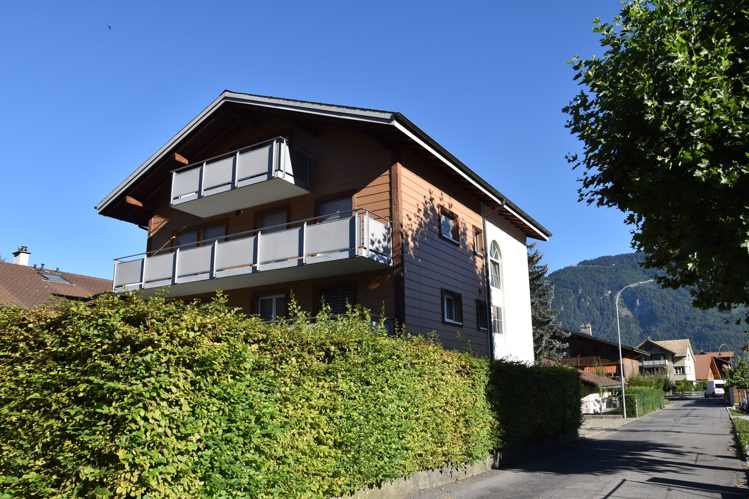 A three-story building with brown and white exterior walls, balconies, and glass windows. Behind the building is a hedge, with mountains in the distance.