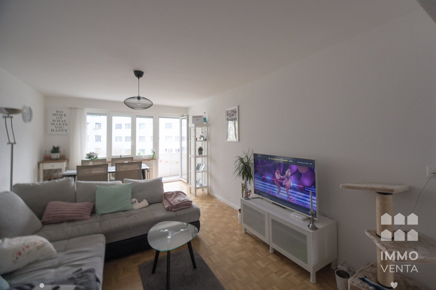 modern living room with gray couch, glass table, TV, wooden floor, white walls, dining table, chairs, plants