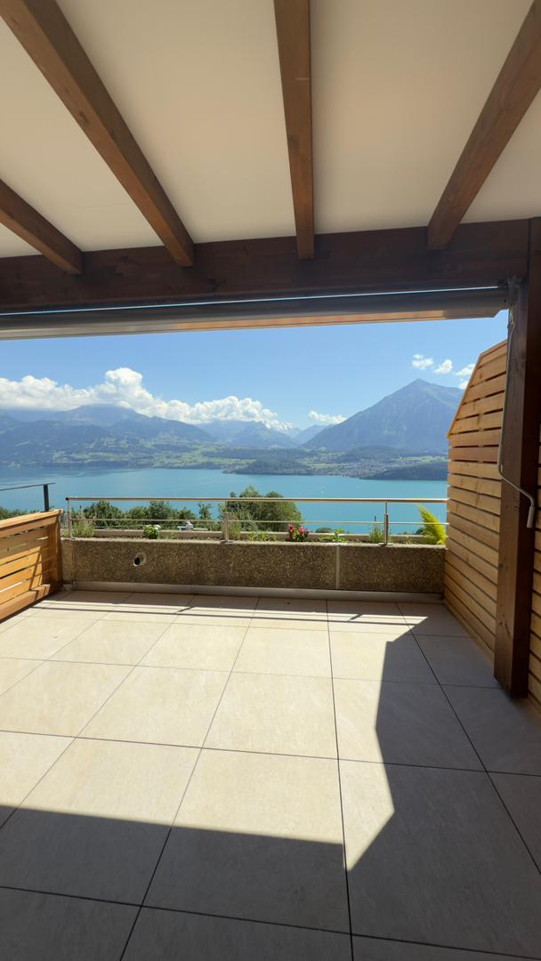 Spacious balcony with a stunning view of a lake surrounded by mountains. The balcony has a tiled floor and wooden beams overhead. There is a railing along the edge of the balcony, providing a safe and unobstructed view of the scenic landscape.