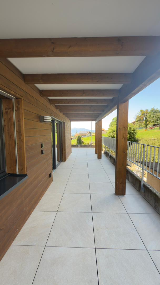 The image shows a covered outdoor walkway or balcony area with a tiled floor and wooden beams supporting the ceiling. The walkway leads to what appears to be a residential building, with large windows and a view of the surrounding countryside visible in t