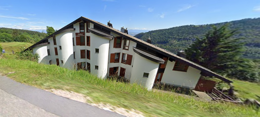 Multi-story apartment building with white exterior walls, brown roofing, and wooden balconies or terraces. The building is situated on a grassy hill with a forested area in the background.