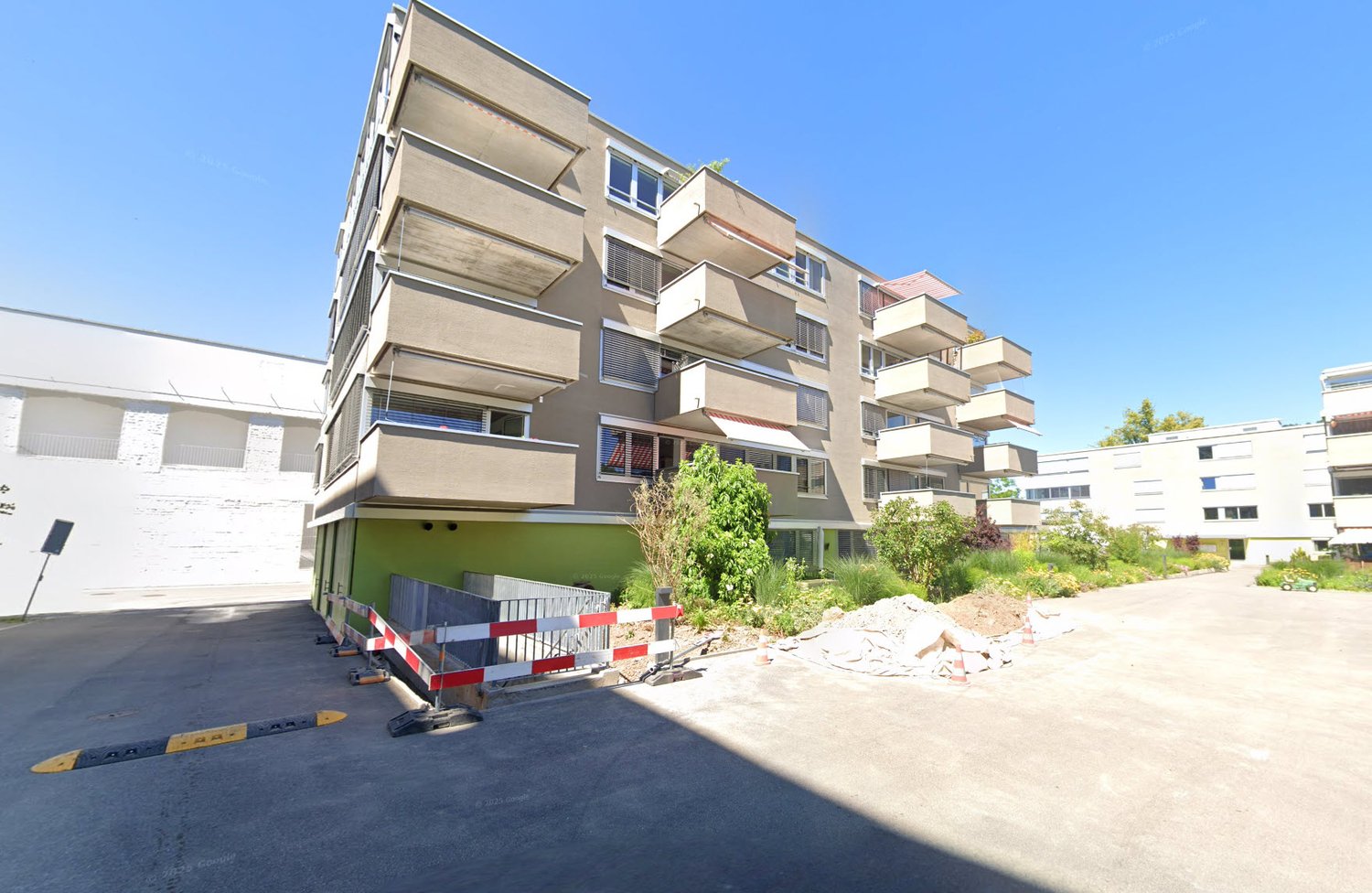 Apartment building, multiple floors, balconies, greenery, entrance area, open parking lot, under construction signs.