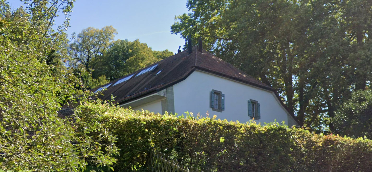White and brown gable roof, two windows, small bushes, large green trees in the background