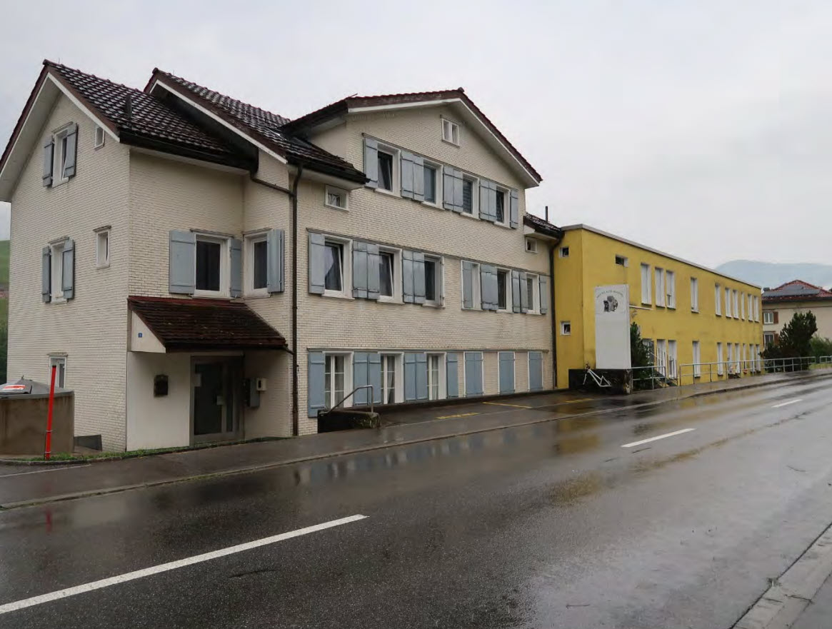Two-story house with brick facade, slate roof, multiple windows with blue shutters, and entrance with an awning.