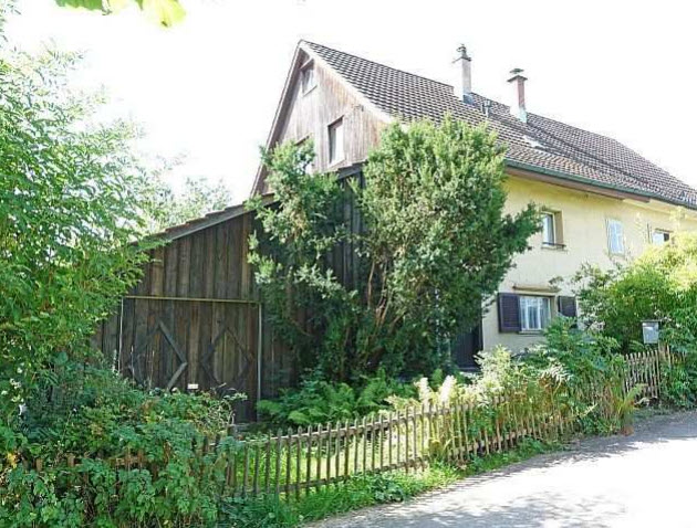 a two story house, yellow color, wooden exterior on the left side, trees and plants surrounding it, small wooden fence in the front, two windows on the right side of the house, two chimneys