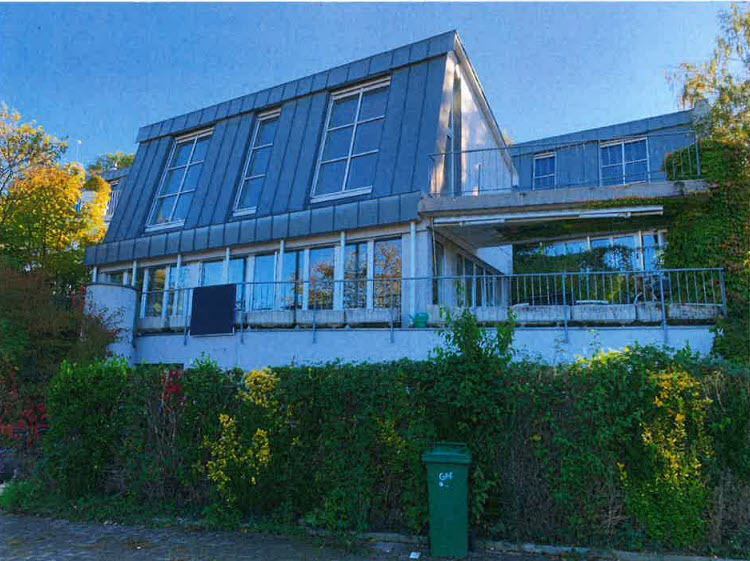modern house, gray walls, multiple large windows, balcony with railing, surrounded by greenery