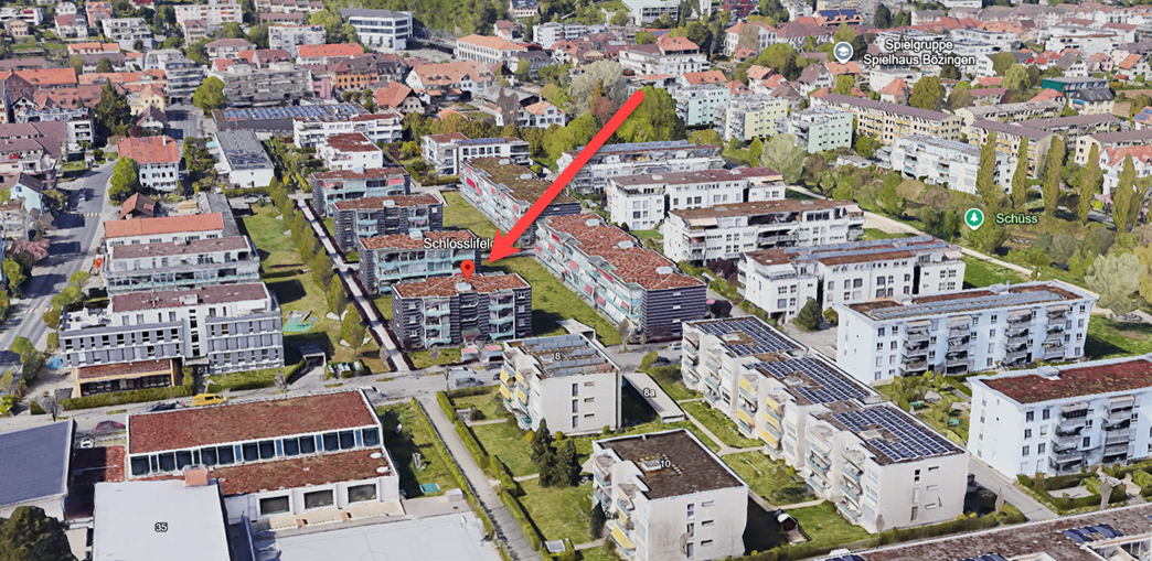 Aerial view of a residential area with multiple buildings, some with solar panels on the roofs, a central green space, and a red arrow pointing to a specific building.
