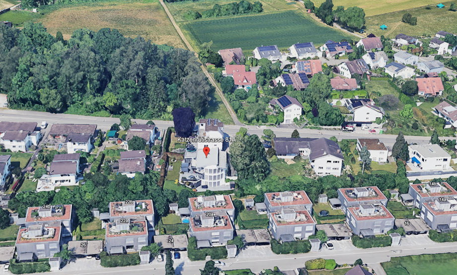 Aerial view of a residential area with multiple houses, some with solar panels, surrounded by trees and greenery, with a central building marked with a red pin.