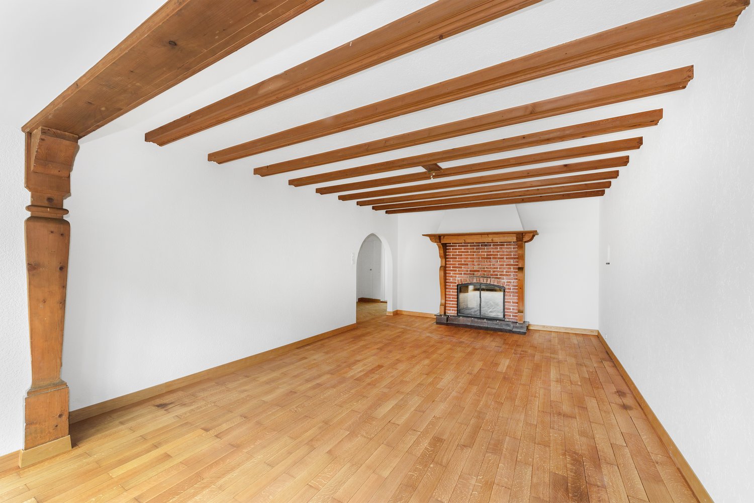 Large open room with exposed wooden beams on the ceiling, hardwood flooring, and a brick fireplace.