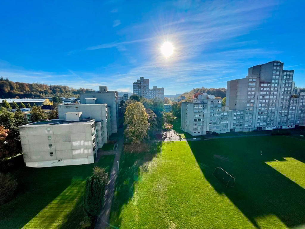 Aerial view of a city skyline with high-rise apartment buildings, a grassy park area in the foreground, and a bright blue sky with a shining sun overhead.