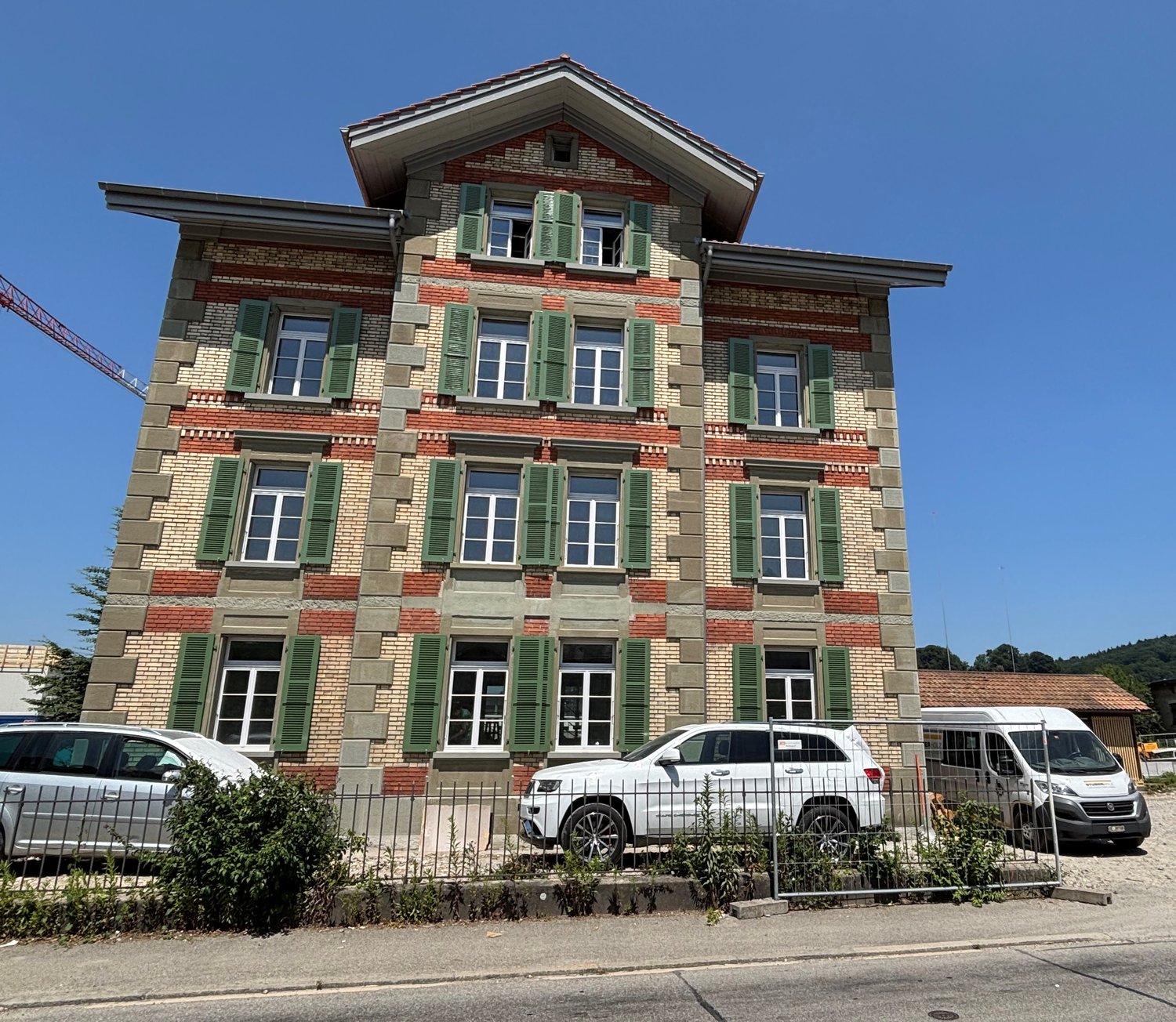 3-story brick and stone building with green shutters, large windows, and a pitched roof. The building appears to be a commercial or residential property, with several vehicles parked in front.