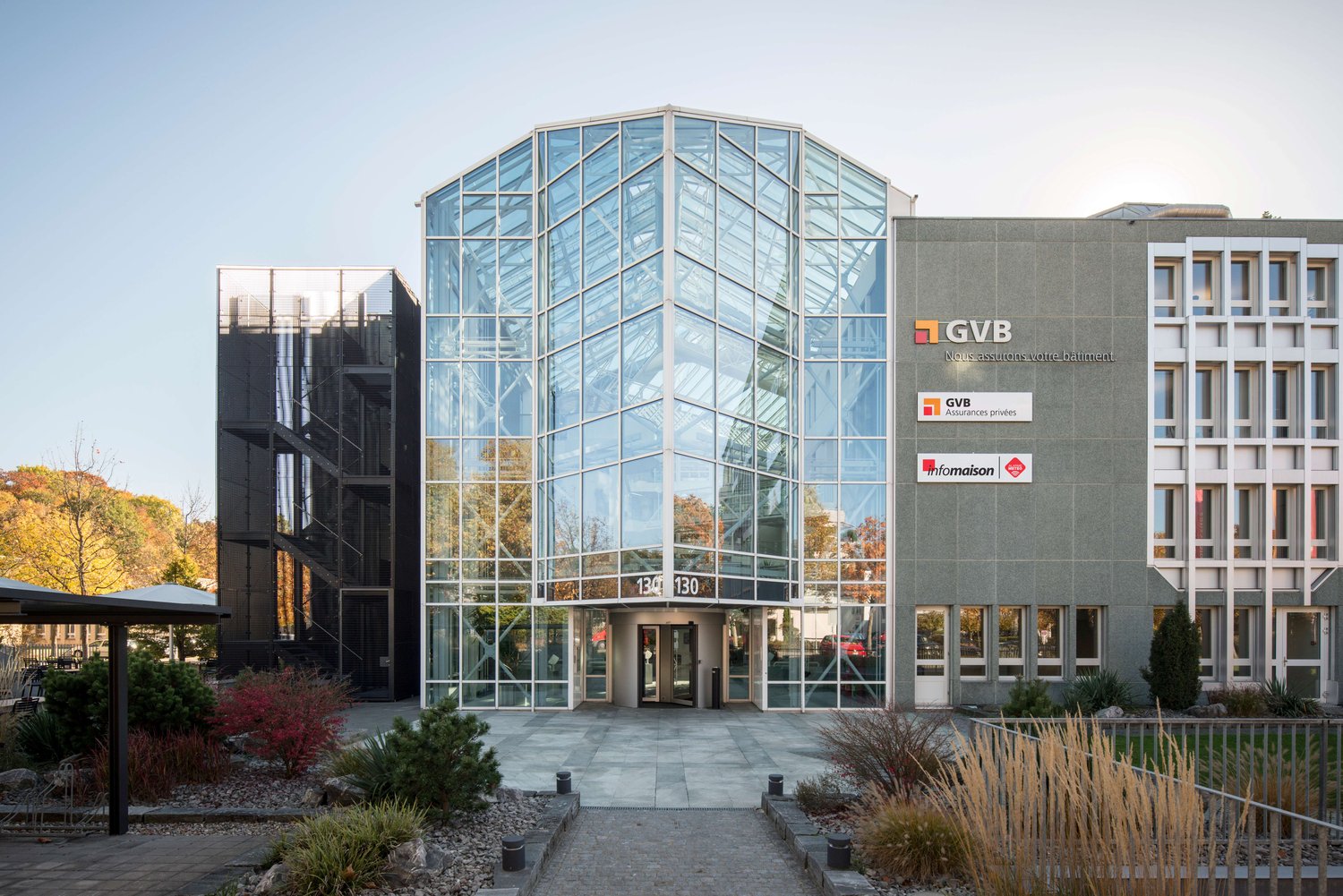 Modern office building with glass facade, entrance with double doors, signage on the wall, surrounded by greenery and a paved walkway.