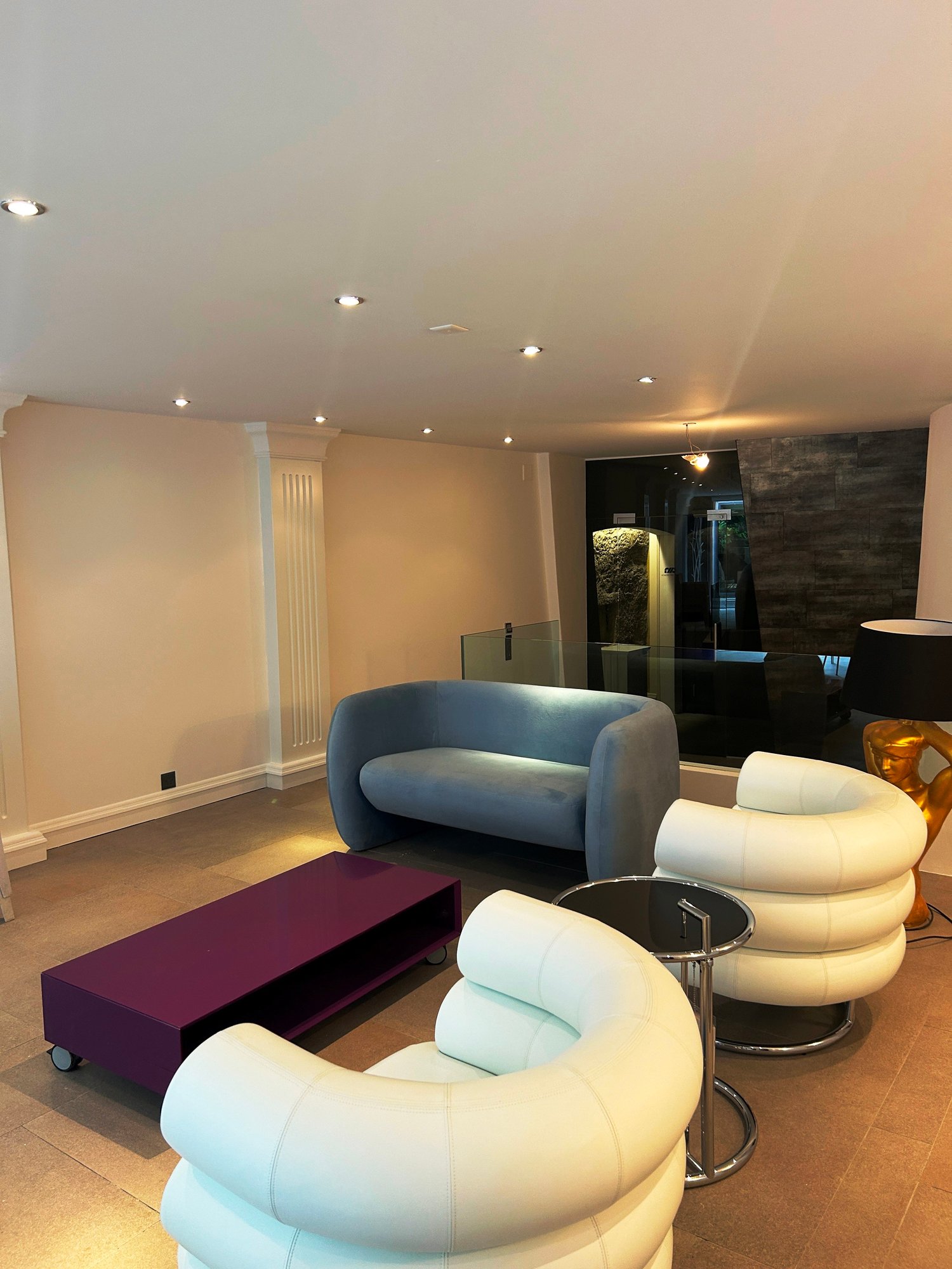 Spacious living room with modern furniture, including a blue sofa, white and beige armchairs, and a purple coffee table on wheels. The room has recessed lighting in the ceiling and a stone accent wall with a fireplace.