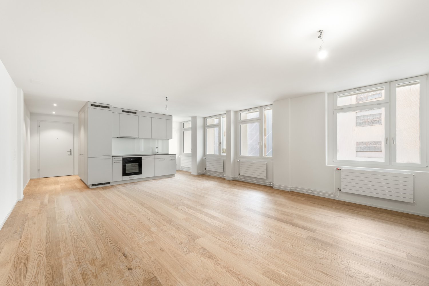 Open layout with kitchen area, large windows, white wooden floors, white walls, built-in appliances