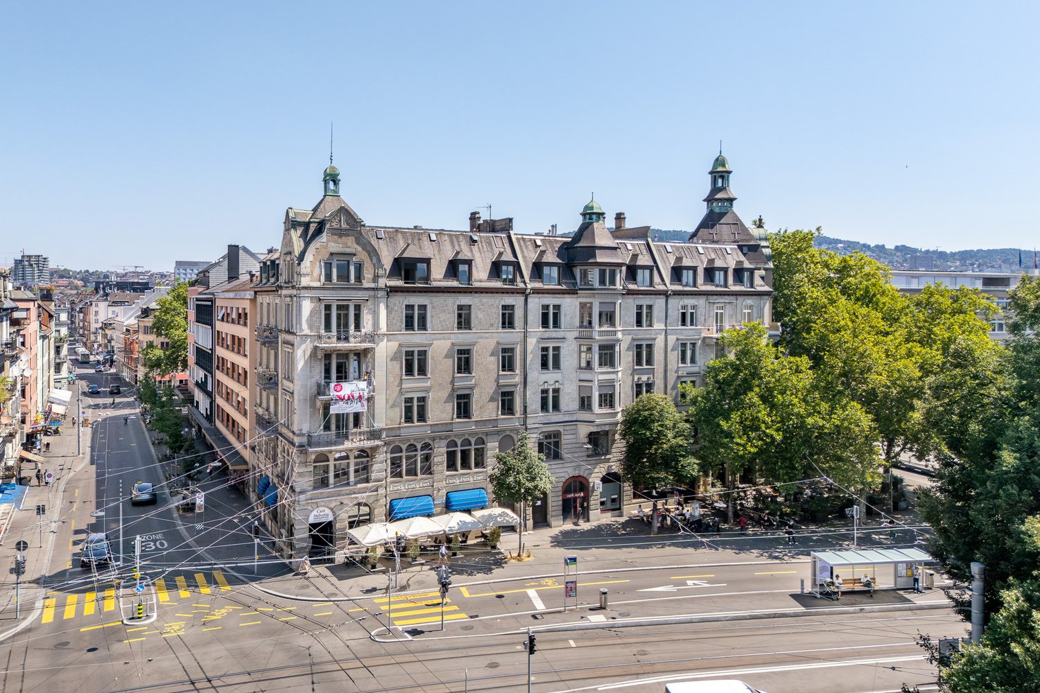 Building with multiple stories, multiple windows, awnings, trees, cars parked in front, an area with outdoor seating and a bus stop