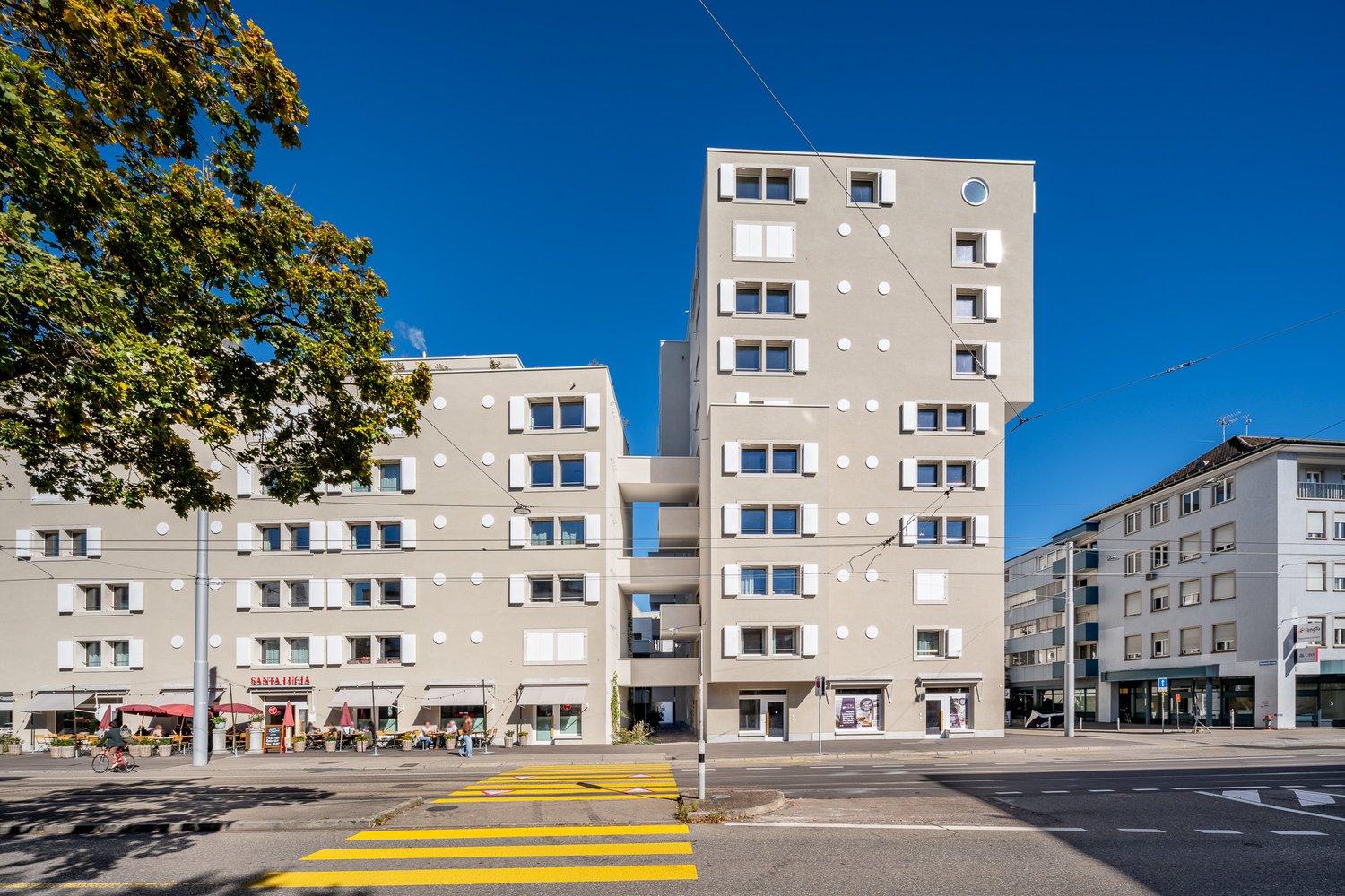 Modern multi-story residential building, beige walls with circular and round windows, connected to smaller buildings with cafes, located on a street corner.