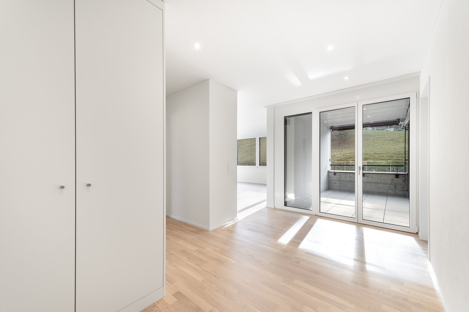 Large room with sliding glass doors leading to balcony, wooden floor, white walls, large windows