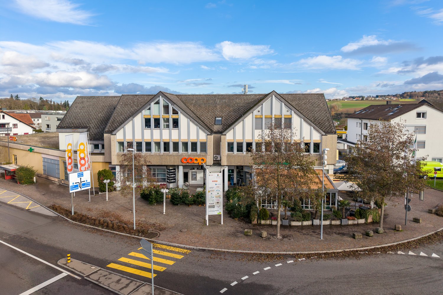 A building with a flat roof, multiple windows, and a sign reading 'COOP'. It is surrounded by trees and plants, with a parking area in front. There are other buildings in the background, and a yellow bus is visible on the right side.