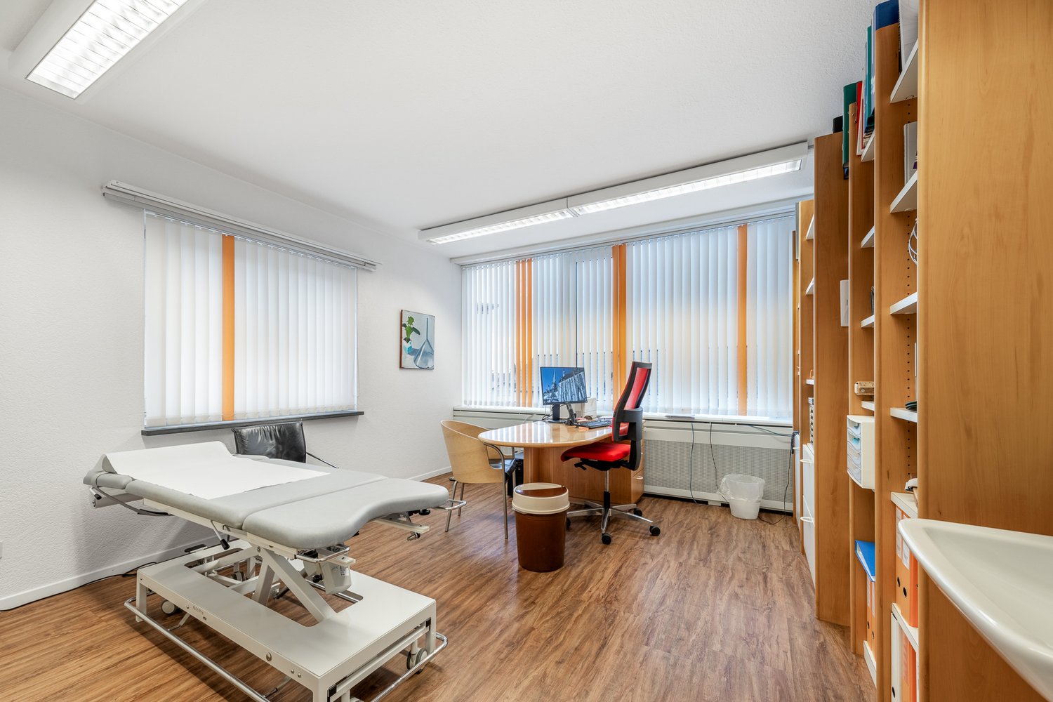 Examination room with examination table, desk, chairs, monitor, radiator, trash bin, shelves, and picture frame.