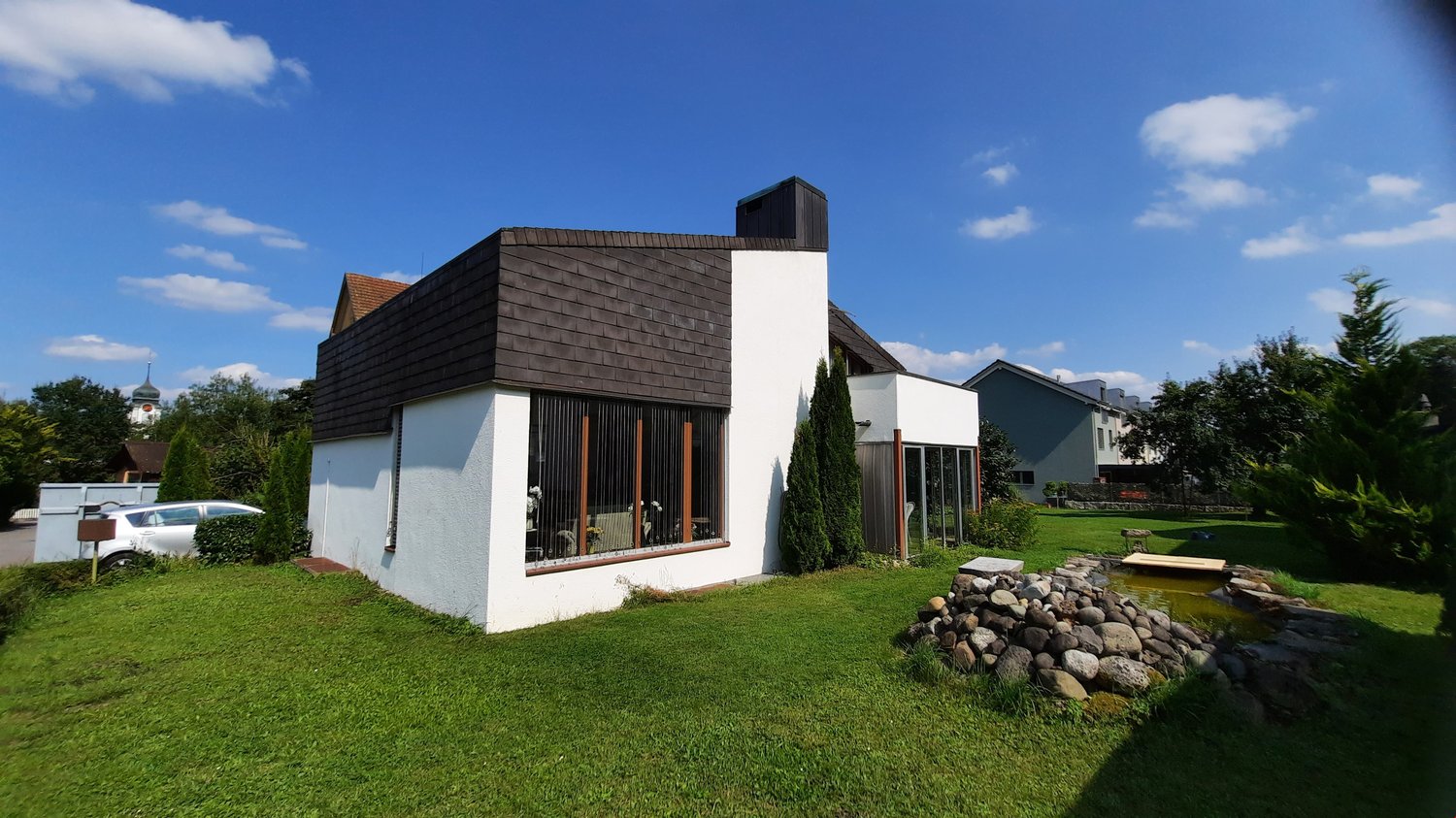 Detached house, white exterior, brown roof, large windows, driveway, parked car, grassy lawn, small pond, shrubs