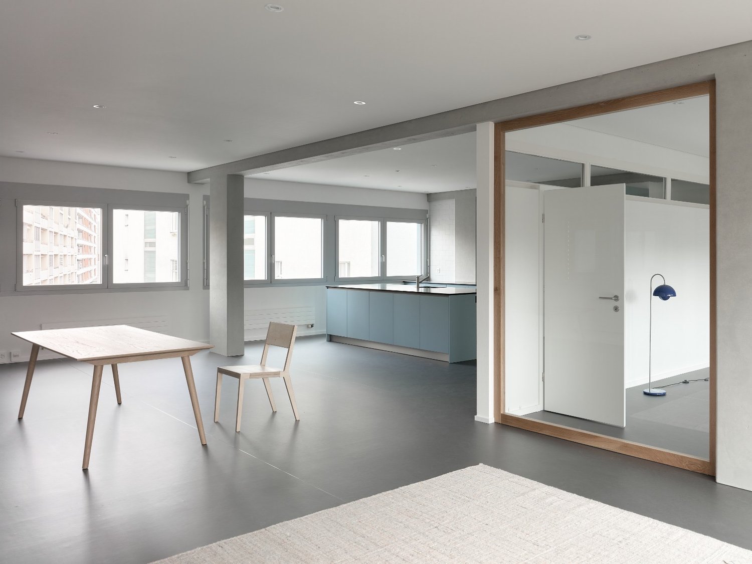 Spacious open-plan living area with a kitchen, dining table, and living room. Large windows provide ample natural light. The kitchen features blue cabinets and a minimalist design. The dining table and chairs are made of wood, complementing the overall mo