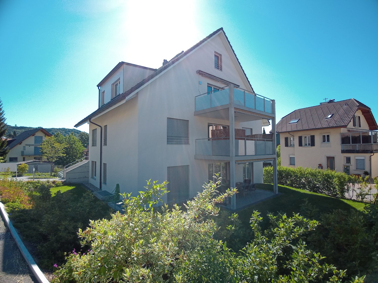 3-story modern house with white exterior, balcony, and surrounded by greenery and other residential buildings