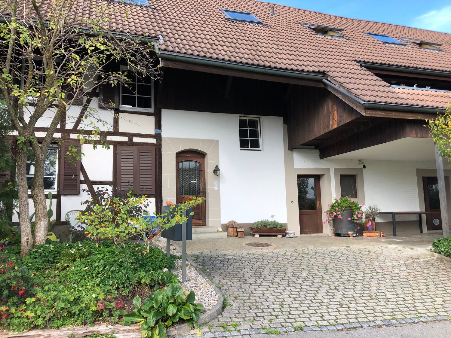 Two-story house with a brown tiled roof, white and brown walls, and wooden shutters, cobblestone driveway, trees, and plants around the property