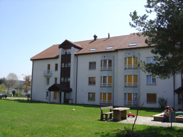 3-story apartment building with white exterior, balconies, and a grassy area in front with benches