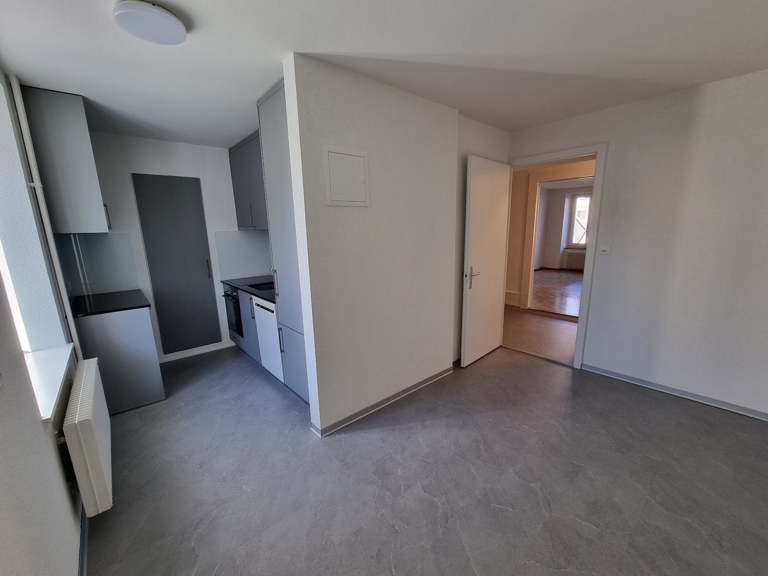 The image shows a studio apartment with a kitchen area, hardwood floors, and a doorway leading to another room. The apartment has a minimalist and modern design with neutral colors and clean lines.