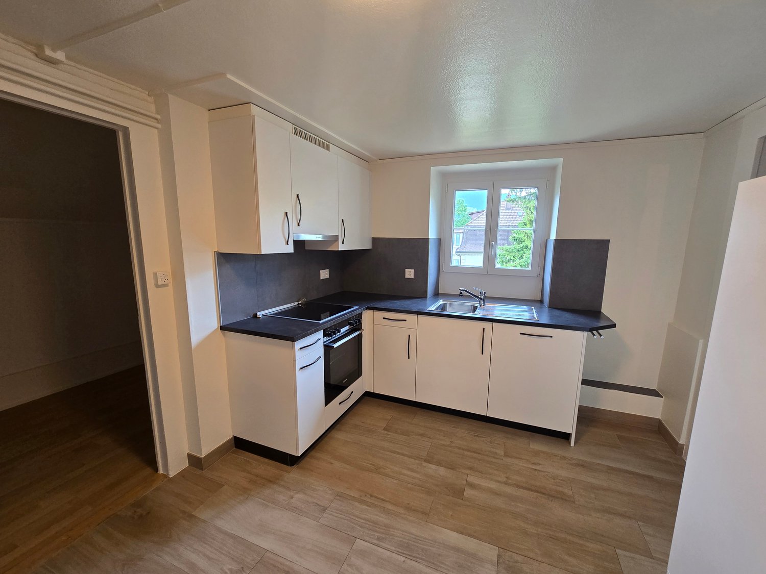 Spacious kitchen with white cabinets, black countertops, stainless steel appliances, and hardwood flooring. The kitchen has an L-shaped layout with a window providing natural light.