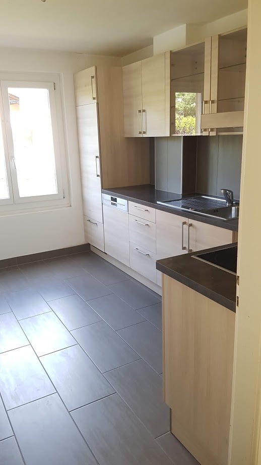 Modern kitchen with light wood cabinets, black countertops, gray tile flooring, and a dishwasher