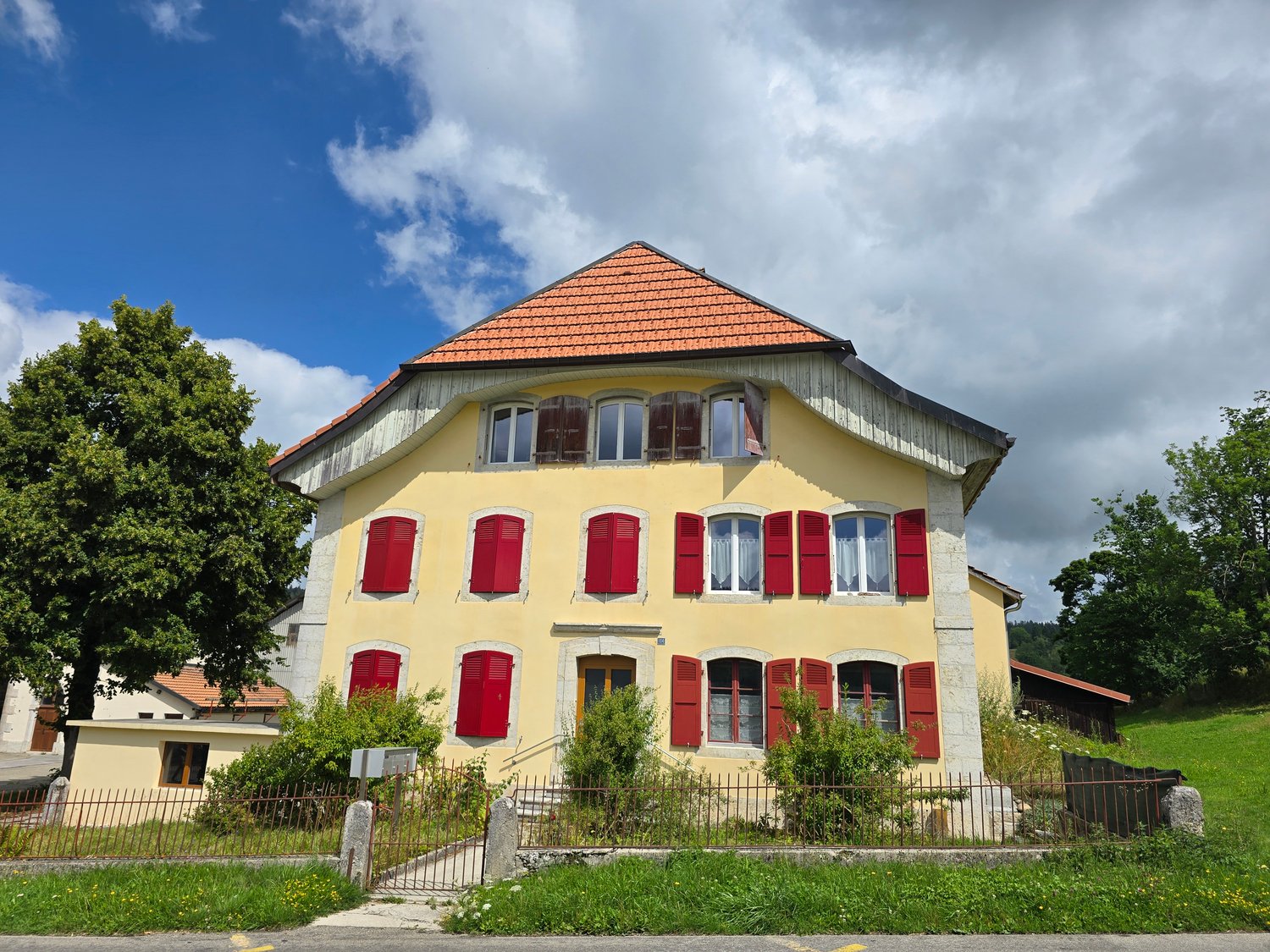 2 story house, yellow exterior, red shutters, tiled roof, surrounded by trees and greenery