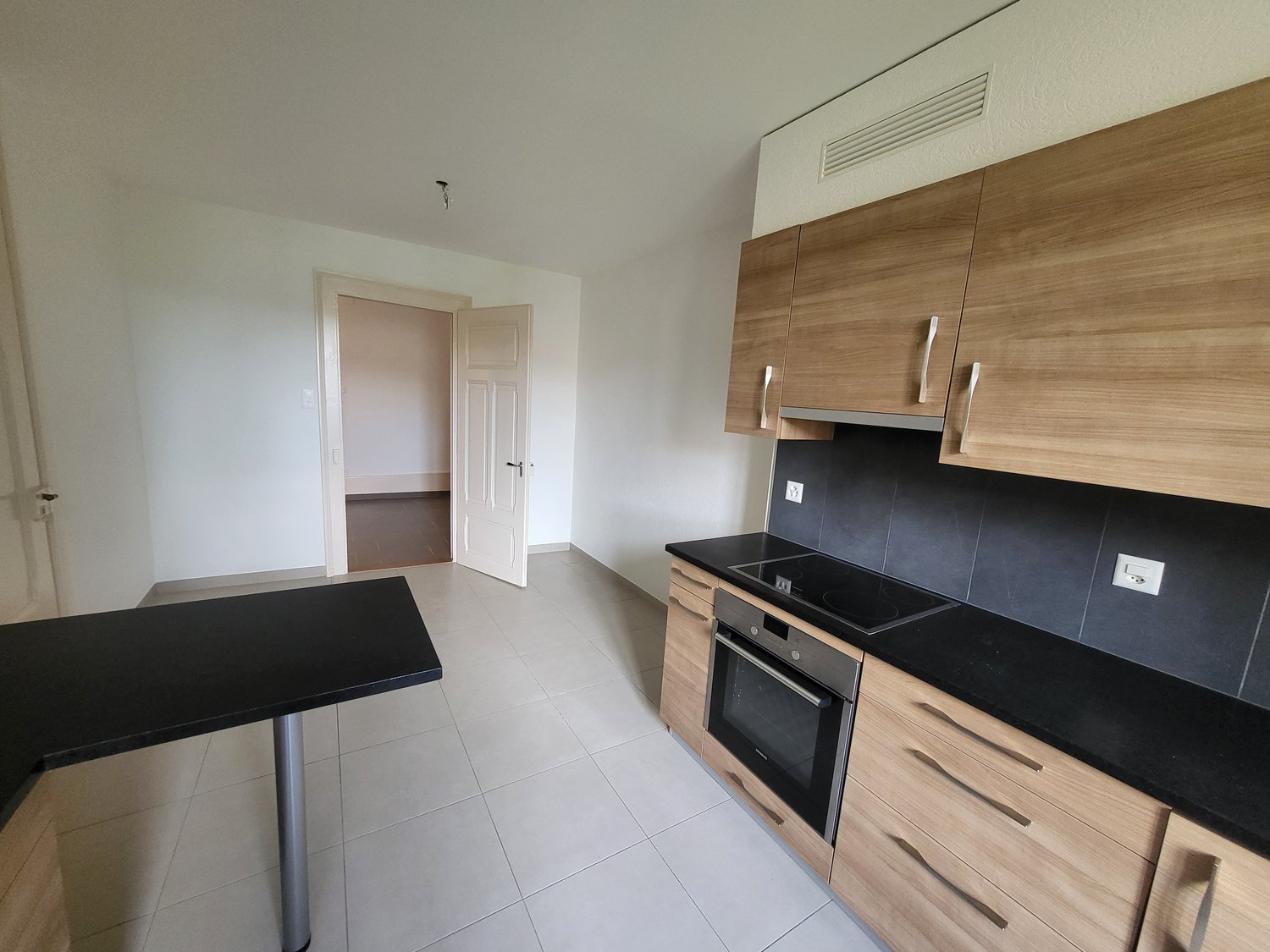 Open plan kitchen with black countertops, wooden cabinets, black stove, and an oven. Tiled floor. Open door leads to another room.