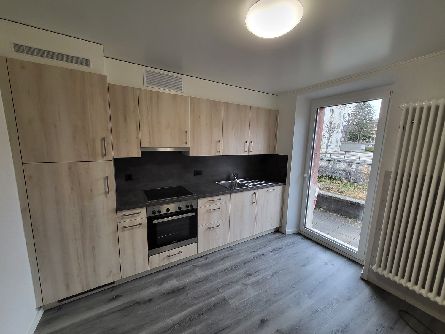 Modern kitchen with wooden cabinets, black countertops, a gas stove, an oven, and a double sink with a faucet. The ceiling has a round light fixture. There's a sliding glass door leading to a paved area with a view of trees and a building.