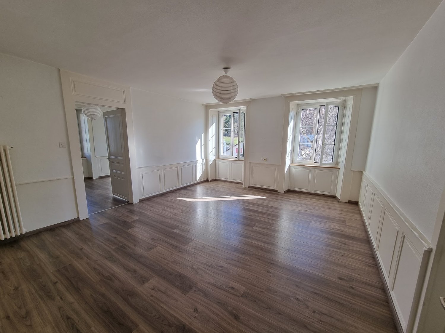 large empty room with wooden floor, white walls, 2 large windows, doors to other rooms