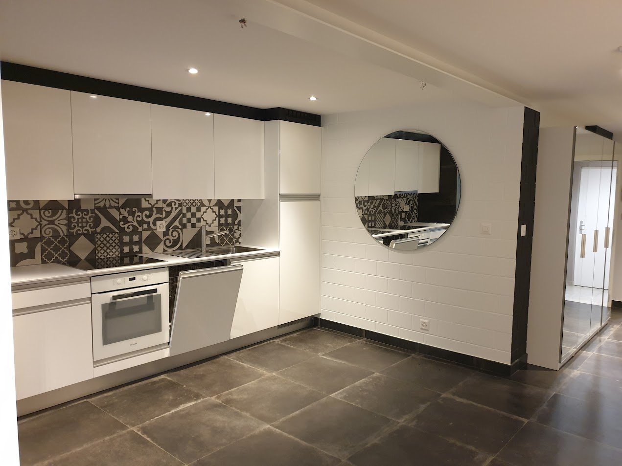 modern kitchen with white cabinets, black tiled backsplash, round mirror, dishwasher, oven, sink, tiled floor