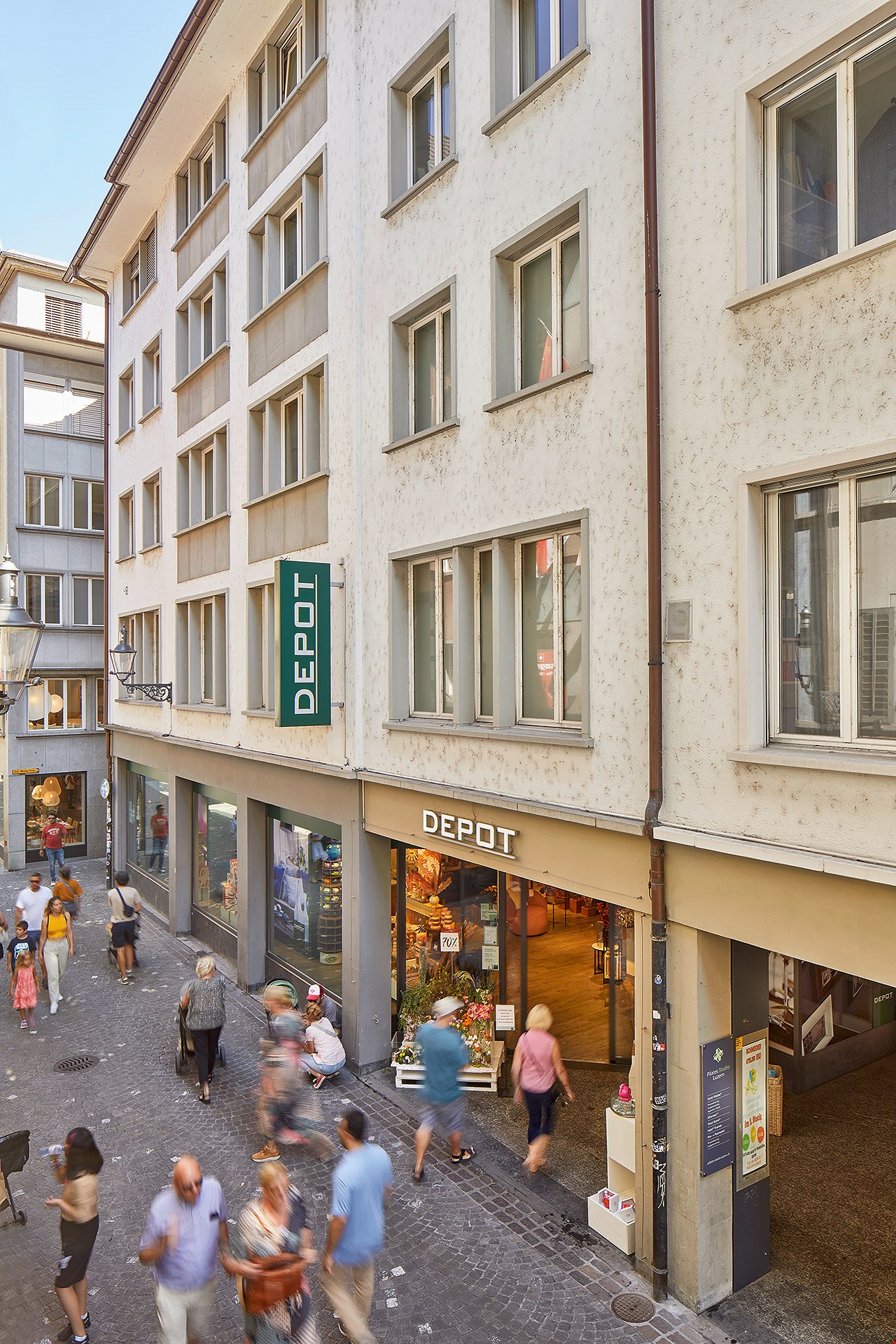 The image shows a street view of a commercial building with a Depot store on the ground floor. The building has multiple floors with windows and a textured exterior. There are people walking on the sidewalk in front of the building.