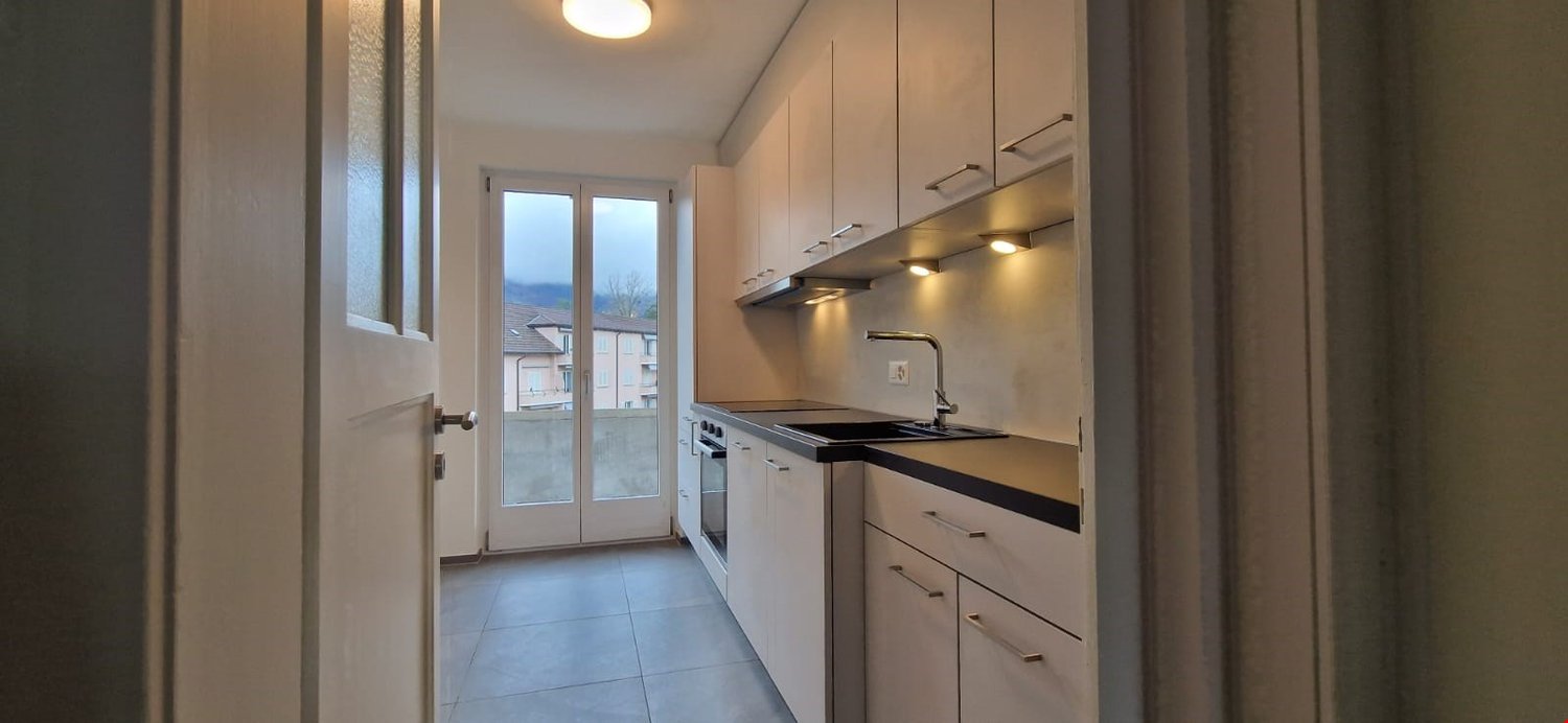 A modern kitchen with white cabinets, a black countertop, a glass door leading to a balcony, and a faucet above the sink