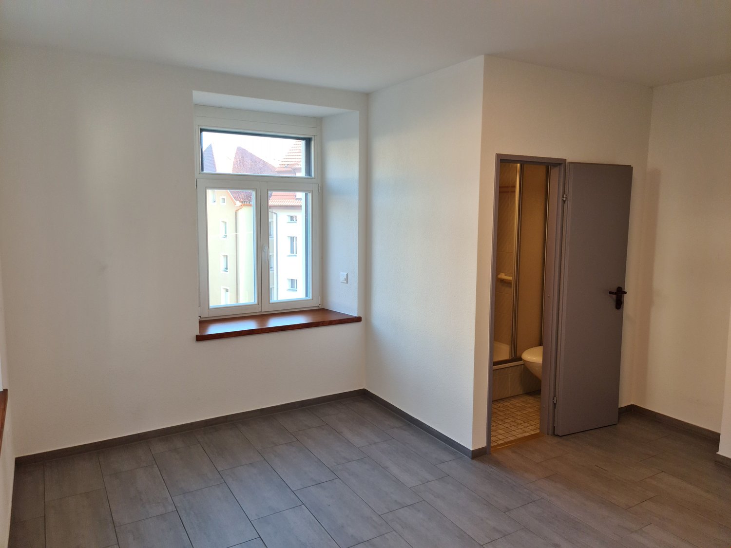 empty room, white walls, wooden tiles, window with window sill, door to bathroom