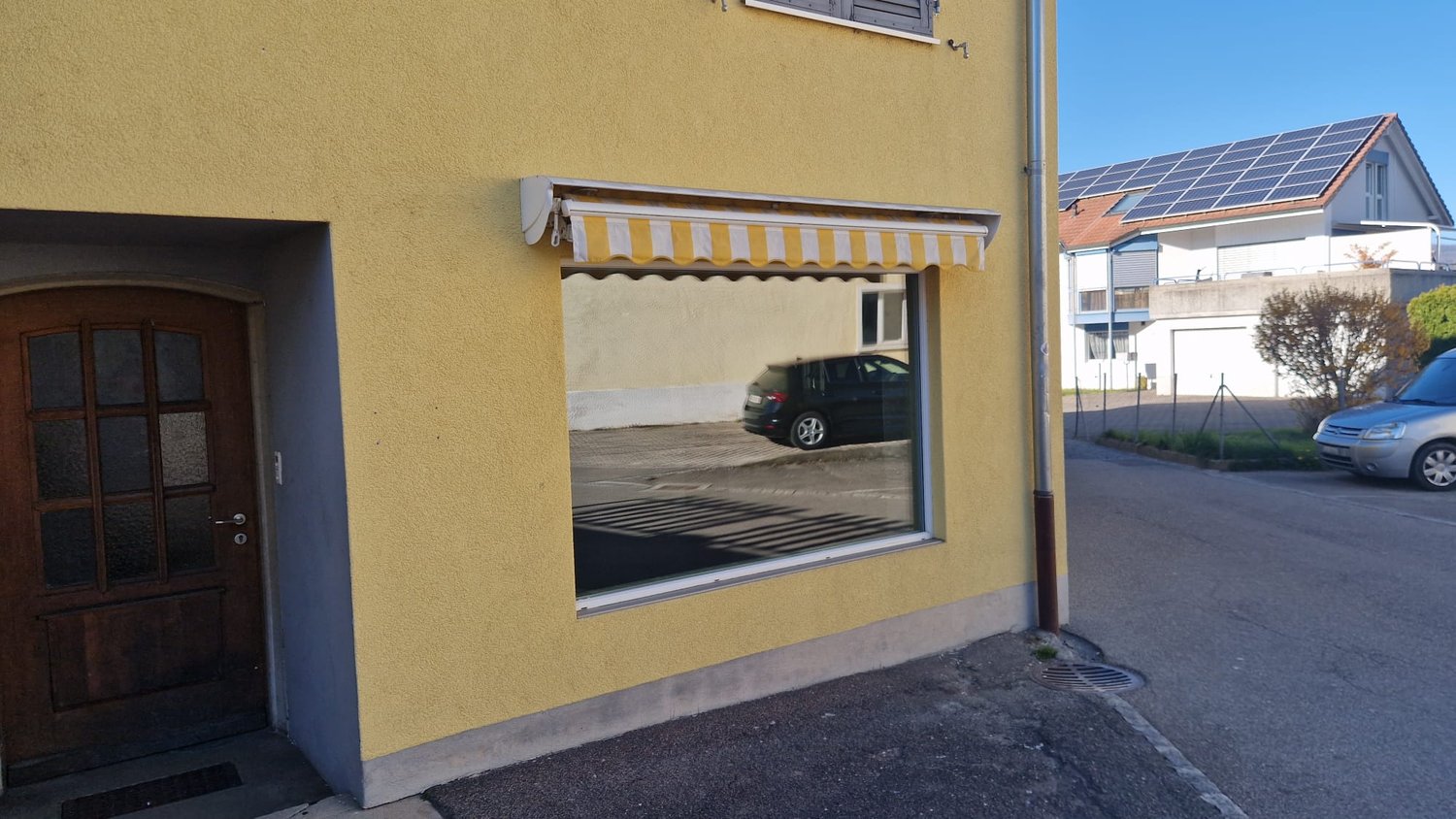 Yellow building with a wooden door, a window with a yellow and white awning, solar panels on the roof, a car parked in front
