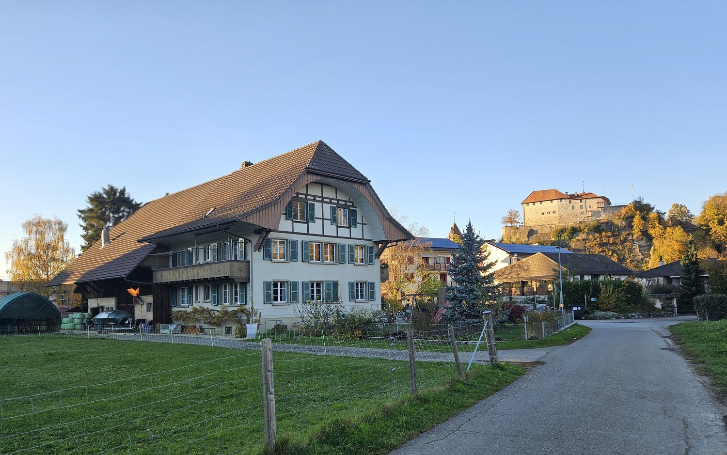 A house with brown roof and white walls, it has many windows, and it is surrounded by trees, plants, and a driveway.