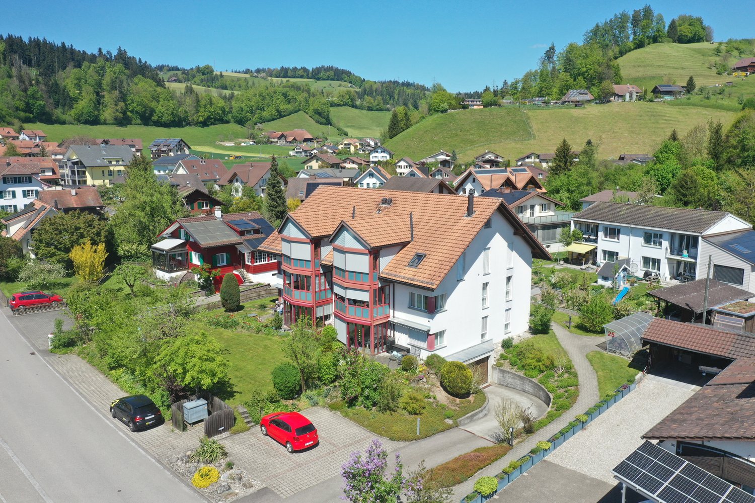Suburban neighborhood with several houses and buildings, surrounded by greenery and hills, featuring a main road, driveways, and cars parked near the houses.