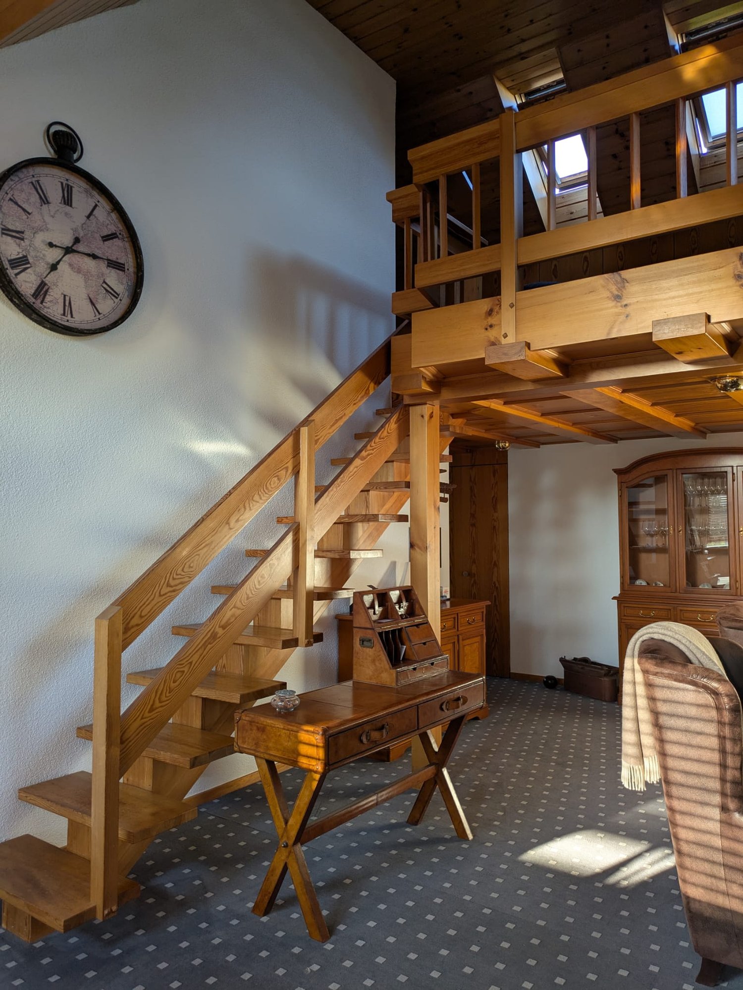 Living room with desk, stairs, wooden cabinets, clock on the wall, carpeted floor, brown couch, brown pillow