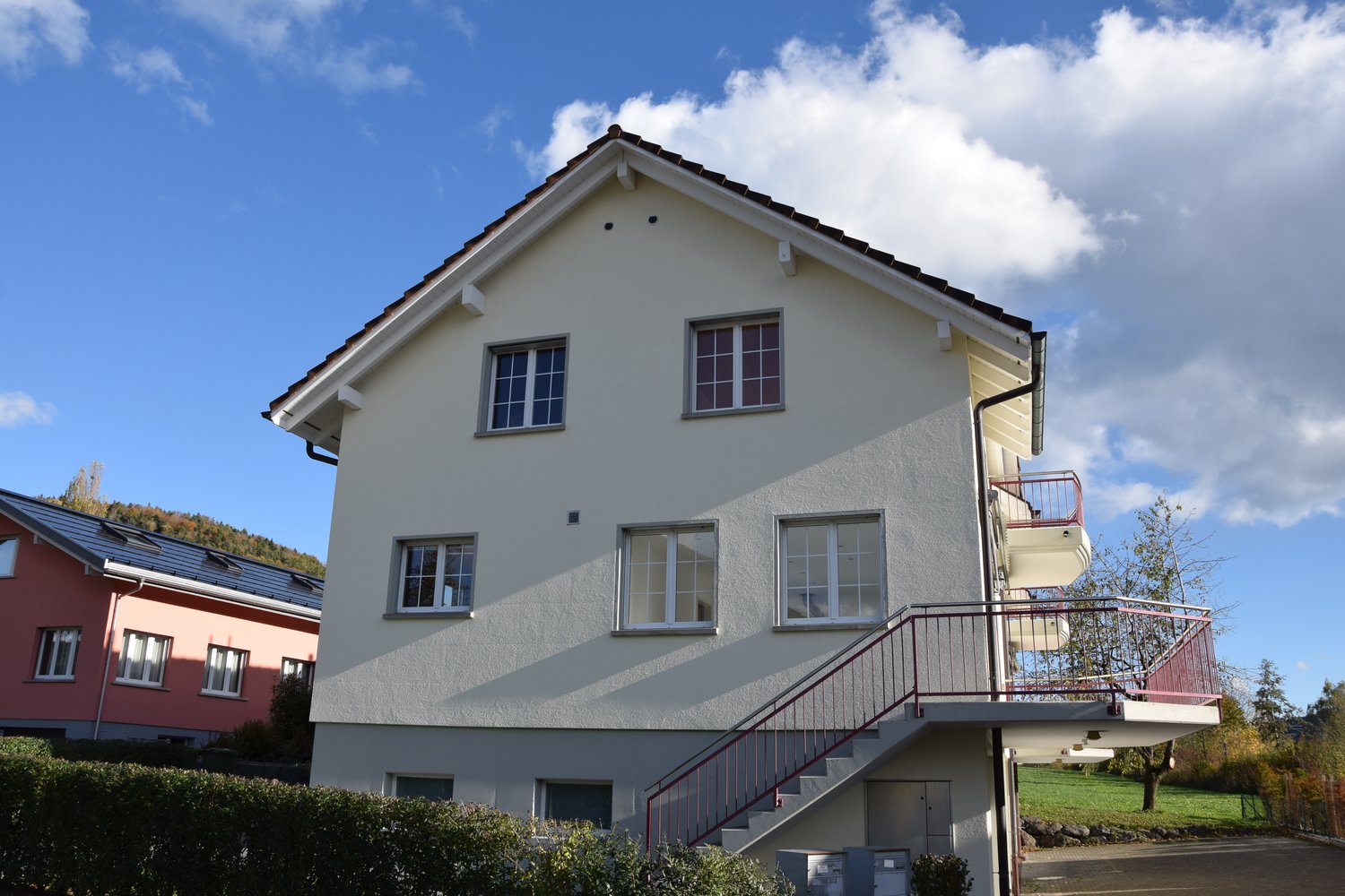 Two-story house, white exterior, red railings on balcony, green bushes in front, gray pathway leading to the entrance.